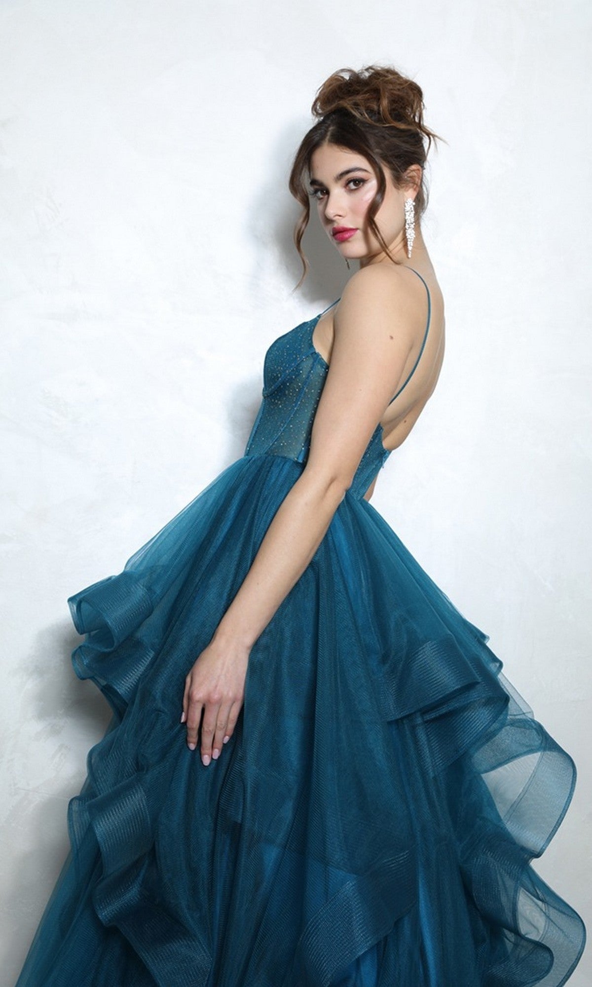 Woman models the Eureka 9950 Long Prom Dress, featuring a blue ballgown style with a ruffled tulle skirt, against a white background. She poses looking over her shoulder, hair up and accessorized with long earrings.